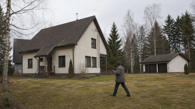 Kuntotarkastaja kiertää muistiinpanovälineiden kanssa 1990-luvun omakotitalon pihalla, taustalla erillinen autotalli.
